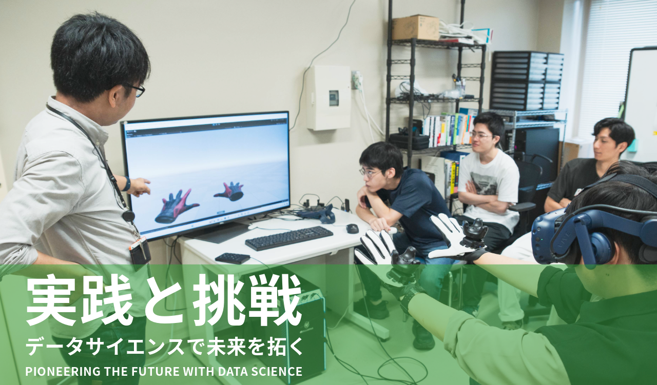 1人の学生がモニターの前に座り、VRゴーグルと手に器具を装着し、モニターにはその学生の手の動きが再現された映像が映し出されている
手前に寺岡助教授がおり、奥にあるモニターを指さしながら数名の学生に説明している様子
